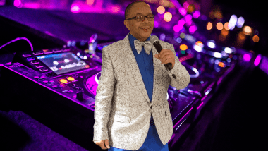 DJ Stan the Man speaking into a microphone with DJ equipment in background. 